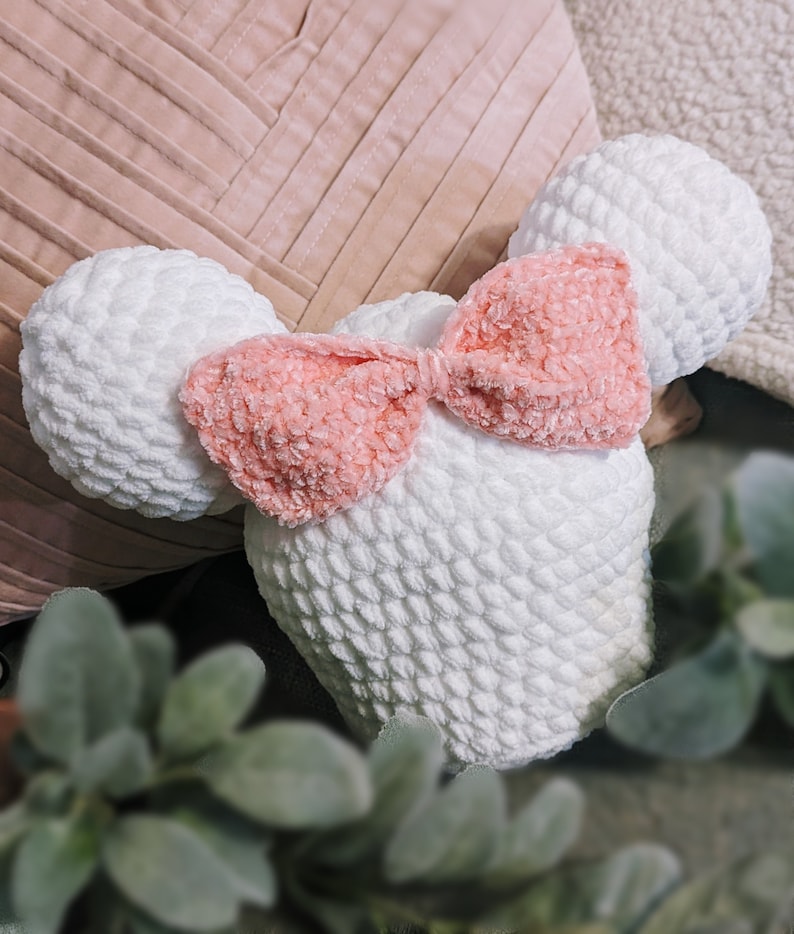 May include: A white crocheted plush toy shaped like a mouse with a pink velvet bow. The toy is sitting on a pink and white striped pillow.