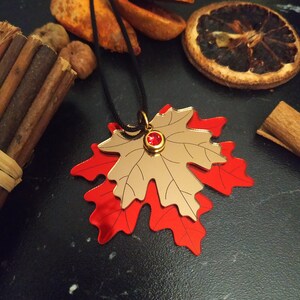 Peut inclure: Un collier pendentif en forme de feuille d'érable superposée, composé d'une feuille supérieure dorée réfléchissante et d'une feuille inférieure rouge vif. Une petite pierre rouge circulaire est sertie dans une monture dorée au centre.
