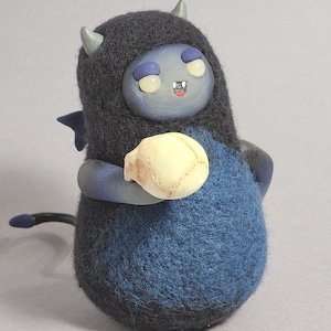 Könnte beinhalten: Eine handgefertigte Filzfigur mit einem dunkelblauen Körper und einer schwarzen Kapuze. Die Kreatur hat kleine graue Hörner, blaue Flügel und hält einen kleinen, cremefarbenen Schädel. Das Gesicht ist blau mit leuchtend gelben Augen und einem kleinen offenen Mund.