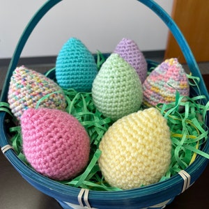 Puede incluir: Una cesta azul llena de huevos de Pascua de crochet en varios colores pastel, incluyendo rosa, amarillo, verde y blanco.