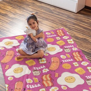 May include: Pink fleece blanket with a breakfast food pattern. The blanket features cartoon images of eggs, bacon, sausage, waffles, and coffee mugs. The name "Tessa" is repeated throughout the blanket.