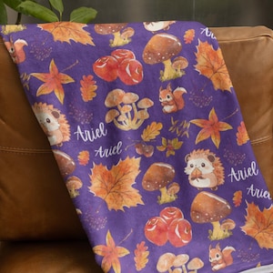 Può includere: Coperta in pile viola con un motivo ripetitivo di funghi, foglie e ricci. La coperta ha il nome "Ariel" stampato più volte.