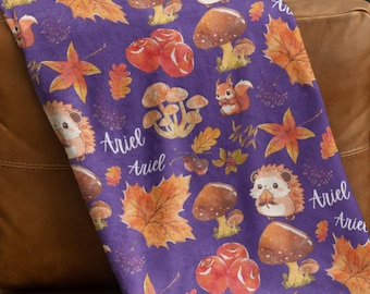 Könnte beinhalten: Lilafarbene Fleece-Decke mit einem sich wiederholenden Muster aus Pilzen, Blättern und Igeln. Die Decke trägt den Namen "Ariel" mehrmals aufgedruckt.