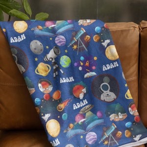 May include: A blue space-themed blanket featuring planets, stars, rockets, telescopes, and an astronaut. The name "ADAM" is printed multiple times across the blanket. The blanket is folded over a brown leather couch.
