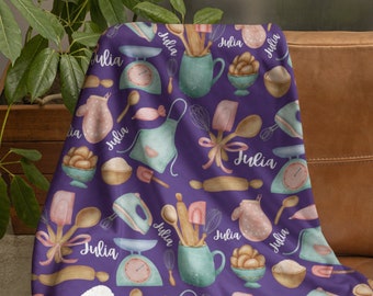Puede incluir: Manta de forro polar morado con forro de sherpa blanco. La manta presenta un patrón repetido de utensilios de cocina y el nombre "Julia" en una fuente rosa claro.
