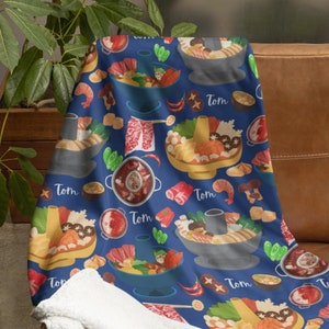 Könnte beinhalten: Eine blaue Decke mit einem Muster aus asiatischen Hot-Pot-Gerichten, Garnelen, Pilzen und Chilischoten. Das Wort "Tom" wiederholt sich im gesamten Design. Die Decke ist über ein braunes Ledersofa drapiert, mit einer weißen, flauschigen Unterseite.