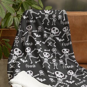 May include: Black Halloween-themed blanket with a white skeleton and bat pattern. The blanket has a soft, white sherpa lining. The name "Frank" is printed on the blanket in a cursive font. The blanket is draped over a brown leather couch.