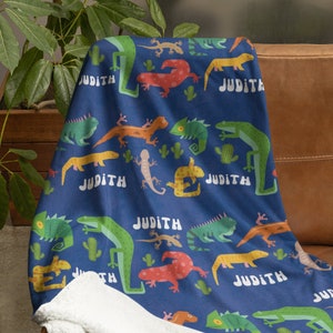 May include: A blue fleece blanket with a white sherpa lining features a repeating pattern of colorful lizards and the name "Judith" printed multiple times.