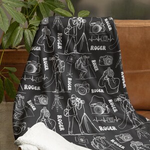 May include: Black and white throw blanket with a repeating pattern of a photographer taking pictures with a camera and tripod. The pattern also includes a heart beat symbol and the name "Roger" repeated throughout.