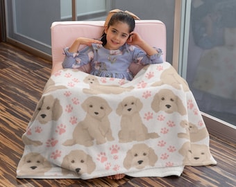 Pode incluir: Um cobertor de lã branca com um padrão de pata rosa e cão doodle dourado. Uma menina está sentada em uma cadeira rosa com o cobertor enrolado ao redor dela.