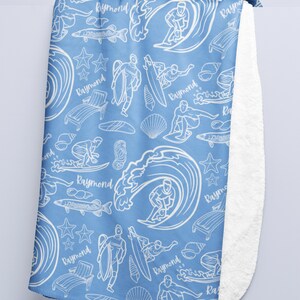 May include: A blue fleece blanket with a white pattern of surfers, waves, fish, seashells, and the name "Raymond" repeated throughout.