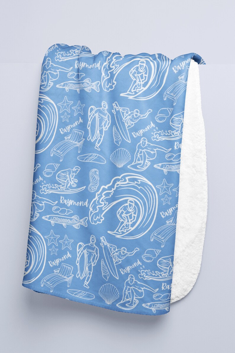 May include: A blue fleece blanket with a white pattern of surfers, waves, fish, seashells, and the name "Raymond" repeated throughout.