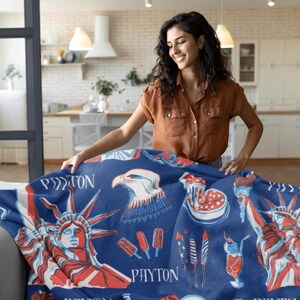 May include: A blue blanket featuring patriotic imagery, including the Statue of Liberty, an eagle, and fireworks. The blanket has red and white accents and the name "PAYTON" printed on it. The blanket is being held by a person.