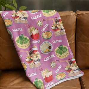 Könnte beinhalten: Eine rosa Fleece-Decke mit einem sich wiederholenden Muster aus asiatischem Essen, darunter Nudeln, Knödel und Reis. Die Decke trägt den Namen "Carter" aufgedruckt.
