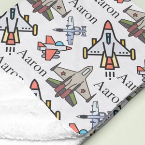 Puede incluir: Manta de bebé blanca con un patrón de avión colorido. La manta presenta diferentes tipos de aviones, incluidos aviones de combate, aviones de hélice y helicópteros. El nombre "Aaron" se repite en todo el diseño.