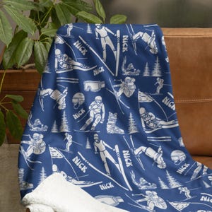 Può includere: Una coperta blu con un motivo bianco a tema sci. Il design include sciatori, sci, occhiali e il nome "Nick" ripetuto. La coperta è drappeggiata su un divano in pelle marrone.