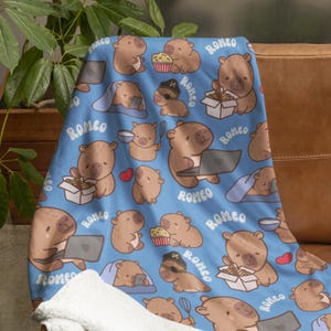 May include: A blue fleece blanket with a repeating pattern of brown capybaras doing various activities. The capybaras are wearing pyjamas, eating popcorn, using laptops, and eating noodles. The text "ROMEO" is repeated throughout the pattern.