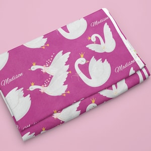 May include: Pink fabric with a white swan pattern. The swans are wearing crowns and have pink flowers. The fabric says "Madison" on it.