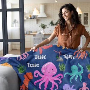 May include: A blue fleece blanket with a colorful octopus pattern. The blanket features a pink octopus with a smiling face, a green octopus, and a red octopus. The blanket also has the name "Teddy" printed on it.