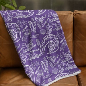 May include: Purple fleece blanket with a white pattern of surfers, waves, fish, and seashells. The pattern repeats the name "Raymond" throughout.