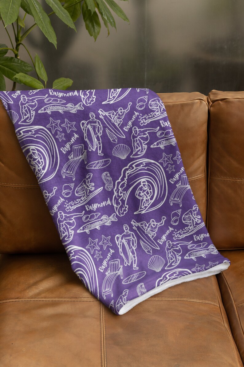 May include: Purple fleece blanket with a white pattern of surfers, waves, fish, and seashells. The pattern repeats the name "Raymond" throughout.