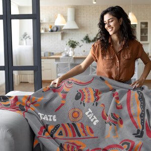 May include: A gray throw blanket with a colorful geometric pattern and the word "Tate" repeated on the blanket. The blanket is being folded on a couch.
