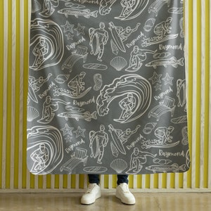 May include: Gray fleece blanket with a white pattern of surfers, waves, fish, and stars. The pattern repeats the name "Raymond" throughout.