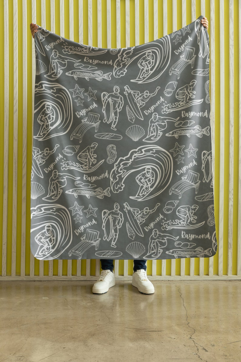 May include: Gray fleece blanket with a white pattern of surfers, waves, fish, and stars. The pattern repeats the name "Raymond" throughout.