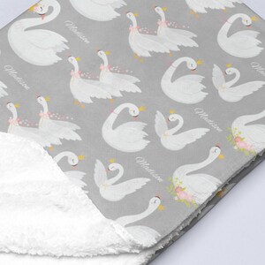 May include: A gray and white baby blanket with a white fluffy backing. The blanket features a repeating pattern of white swans with pink flowers and the name "Madison" printed on the blanket.