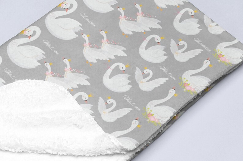 May include: A gray and white baby blanket with a white fluffy backing. The blanket features a repeating pattern of white swans with pink flowers and the name "Madison" printed on the blanket.