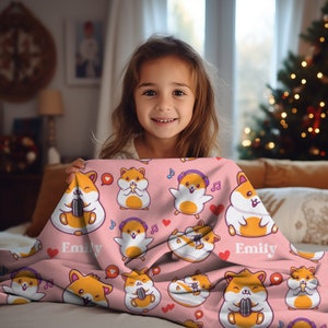 May include: Pink fleece blanket with a repeating pattern of cartoon hamsters wearing headphones and holding microphones. The blanket has the name "Emily" written on it twice.