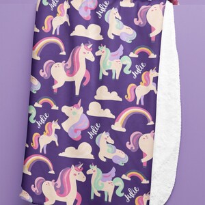 May include: A purple blanket featuring a pattern of unicorns, rainbows, and clouds. The unicorns have pink, purple, and mint green manes and tails. The name "Julie" is written in cursive. The blanket has a white, fluffy underside.