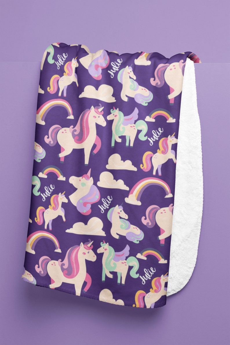 May include: A purple blanket featuring a pattern of unicorns, rainbows, and clouds. The unicorns have pink, purple, and mint green manes and tails. The name "Julie" is written in cursive. The blanket has a white, fluffy underside.
