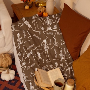 May include: A brown blanket with a white skeleton pattern and the word "Morris" repeated throughout. The blanket is draped over a white couch, with orange and brown pillows. A book, a cup of tea, and decorative pumpkins are also visible.