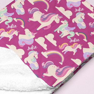 May include: A magenta blanket featuring a pattern of unicorns, rainbows, and clouds. The unicorns have colourful manes and tails. The name "Julie" is printed on the blanket. The underside of the blanket is white.
