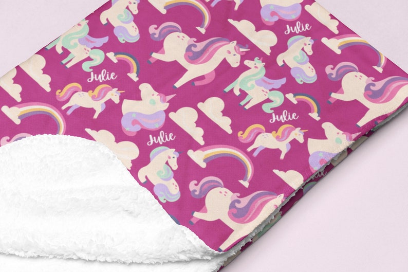 May include: A magenta blanket featuring a pattern of unicorns, rainbows, and clouds. The unicorns have colourful manes and tails. The name "Julie" is printed on the blanket. The underside of the blanket is white.