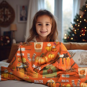 May include: A personalized orange and brown fleece blanket with a fall theme. The blanket features pumpkins, mugs of hot chocolate, and the name "Nate" repeated throughout.