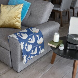 May include: A gray couch with a blue and white throw blanket featuring a swan design and the name "Madison" repeated. The blanket is folded over the arm of the couch. There is a yellow pillow and a blue pillow on the couch.
