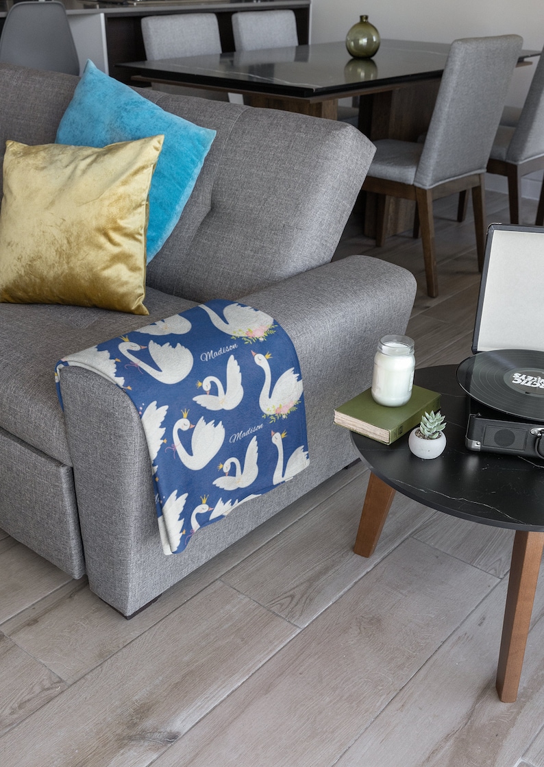 May include: A gray couch with a blue and white throw blanket featuring a swan design and the name "Madison" repeated. The blanket is folded over the arm of the couch. There is a yellow pillow and a blue pillow on the couch.