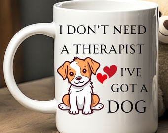 Pode incluir: Caneca de café de cerâmica branca com uma ilustração em preto e branco de um cão e o texto "Não preciso de um terapeuta, tenho um cão" com dois corações vermelhos.