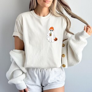 May include: White t-shirt with a graphic of a ghost wearing an orange beanie and holding a coffee cup. The ghost is holding a small orange pumpkin.