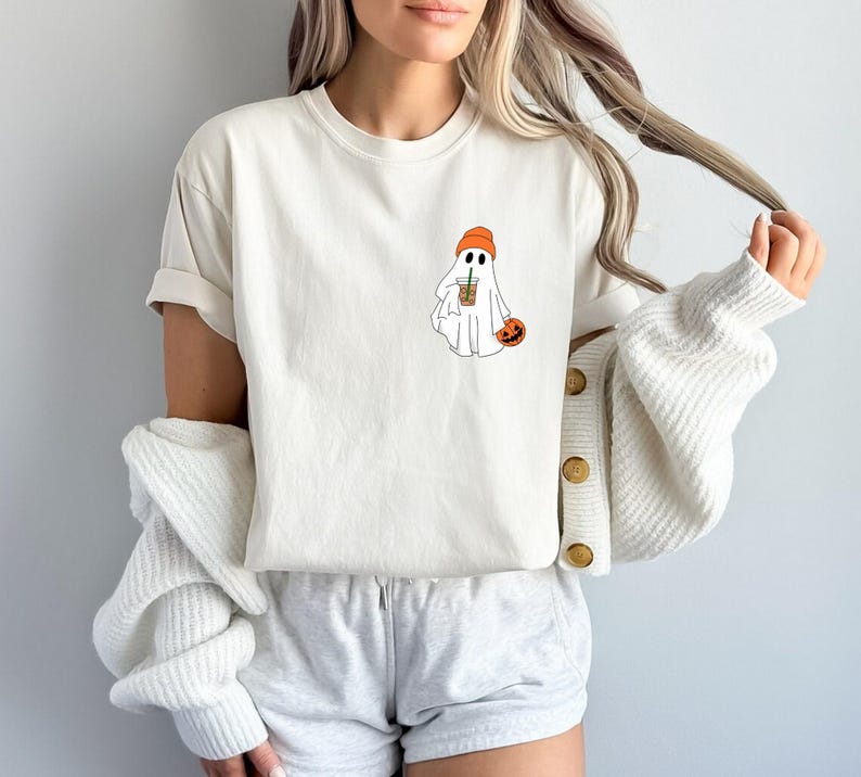 May include: White t-shirt with a graphic of a ghost wearing an orange beanie and holding a coffee cup. The ghost is holding a small orange pumpkin.