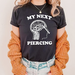 May include: Black t-shirt with white text that reads "My Next Piercing" and a graphic of a brain with a needle going through it.