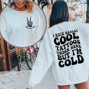 May include: White sweatshirt with a black peace sign and the text "Cool Tattoos" on the front and the text "I have really cool tattoos under here but I'm cold" on the back.