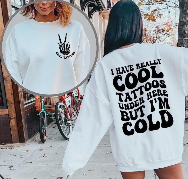 May include: White sweatshirt with a black peace sign and the text "Cool Tattoos" on the front and the text "I have really cool tattoos under here but I'm cold" on the back.