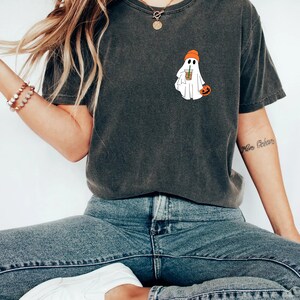 May include: A dark gray t-shirt with a white ghost wearing an orange hat and holding a cup of coffee. The ghost is also holding a small orange pumpkin.