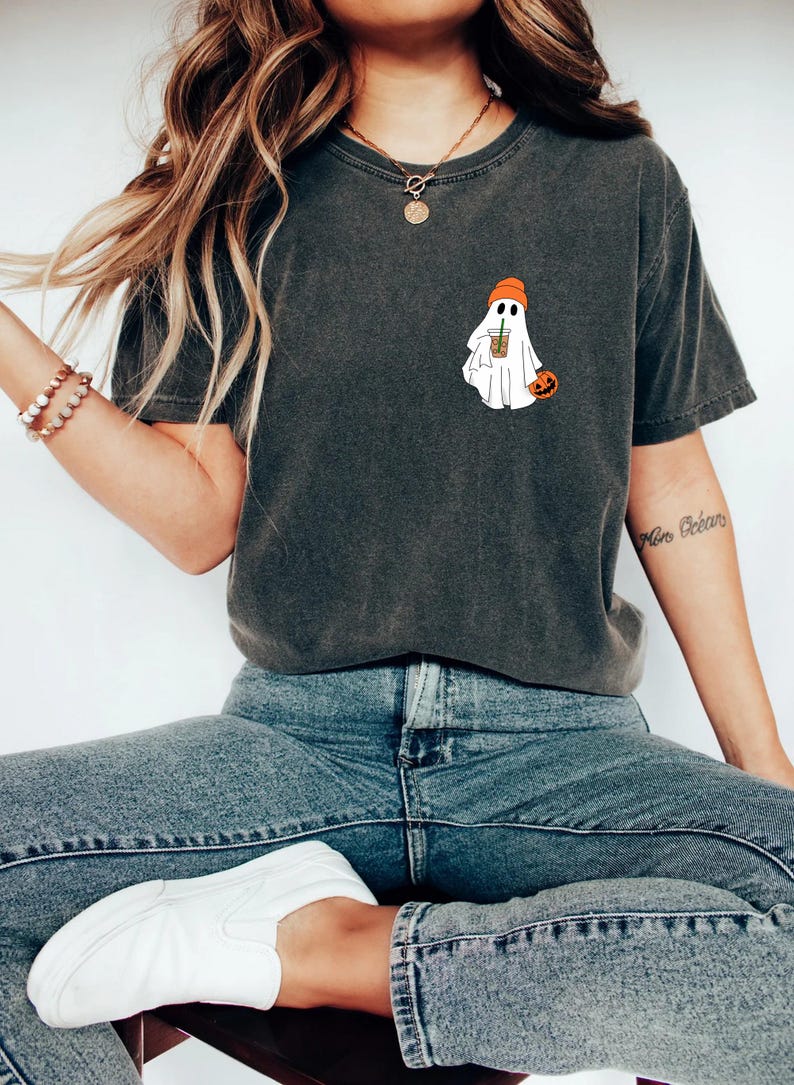 May include: A dark gray t-shirt with a white ghost wearing an orange hat and holding a cup of coffee. The ghost is also holding a small orange pumpkin.