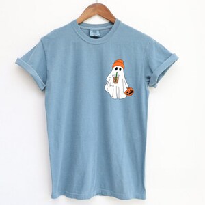 May include: A light blue t-shirt with a white ghost wearing an orange hat and holding a pumpkin and a cup of coffee.