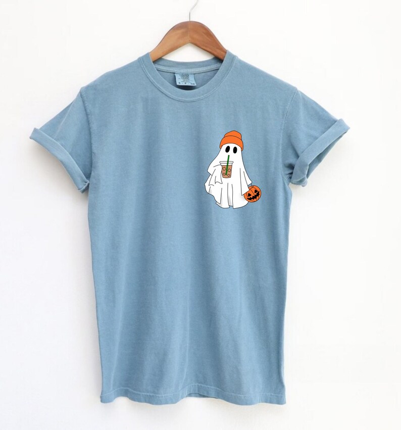 May include: A light blue t-shirt with a white ghost wearing an orange hat and holding a pumpkin and a cup of coffee.