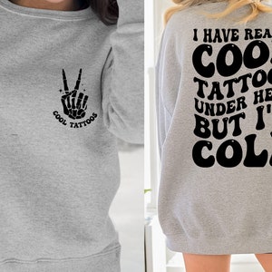 May include: Gray sweatshirt with a peace sign skeleton hand graphic on the left side and the text "I have really cool tattoos under here but I'm cold" on the back.
